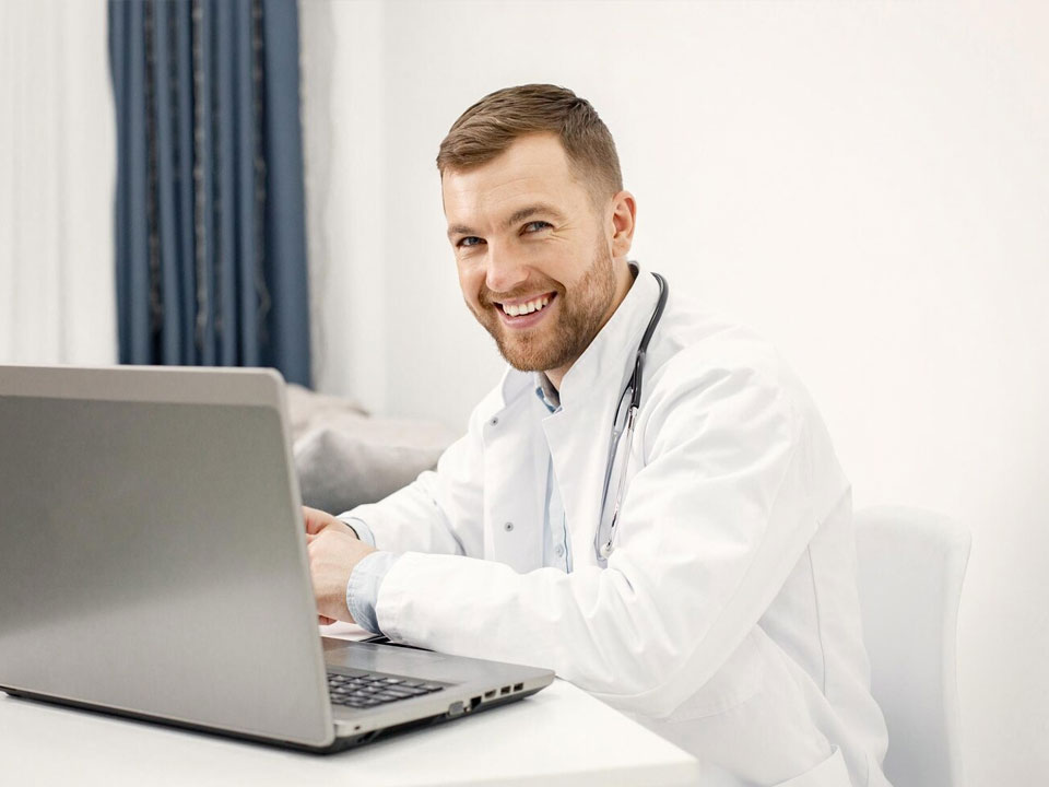 Médico sonriente utilizando tecnología médica en su computadora portátil, representando el uso eficiente de herramientas digitales en la gestión de consultorios modernos.  